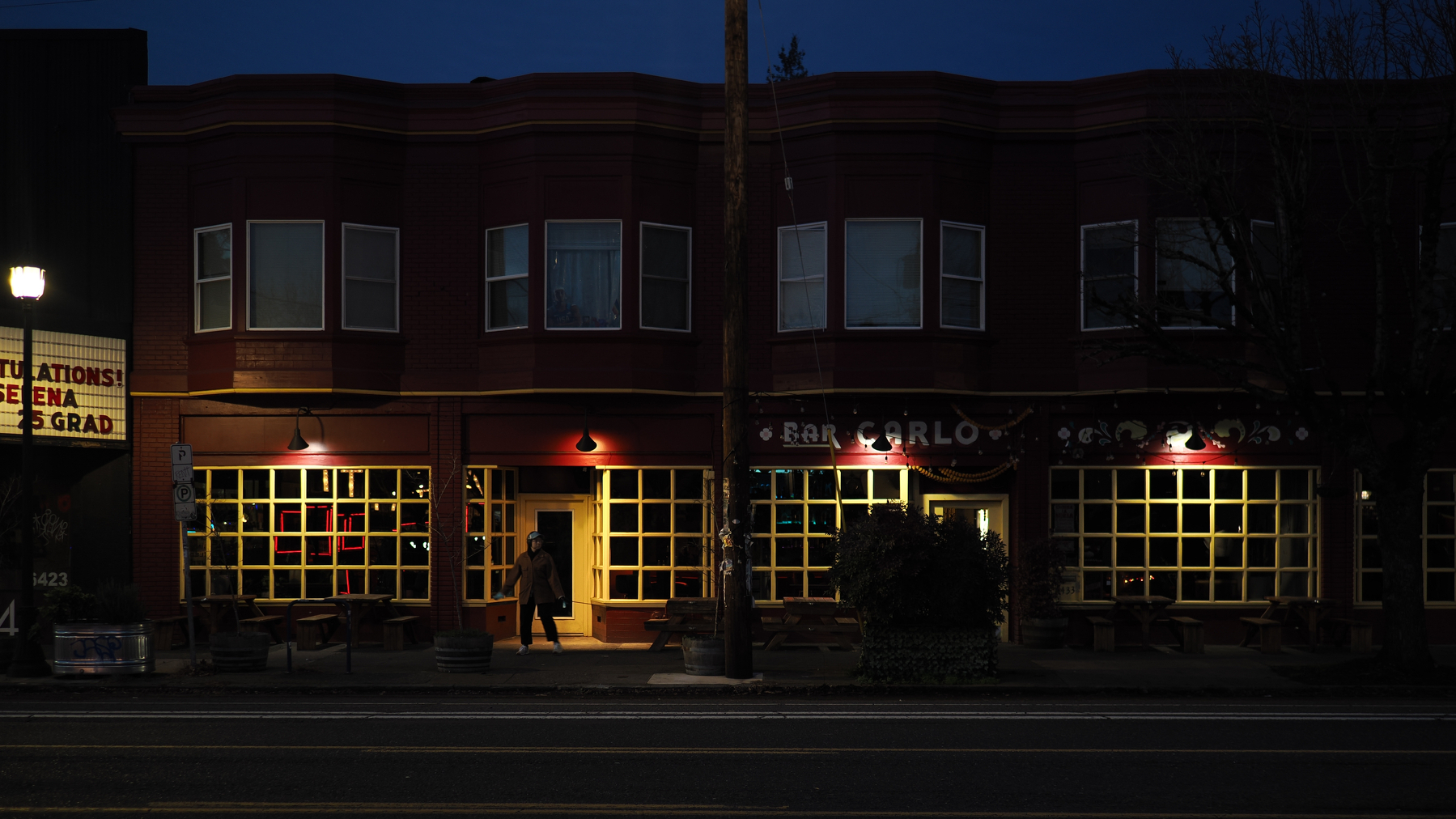 A person walks past a dimly lit storefront with large windows on a quiet street at night.