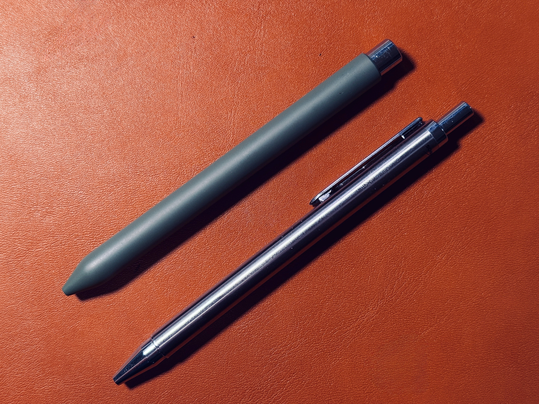 A green pen and a silver pen are placed side by side on a reddish-brown surface.