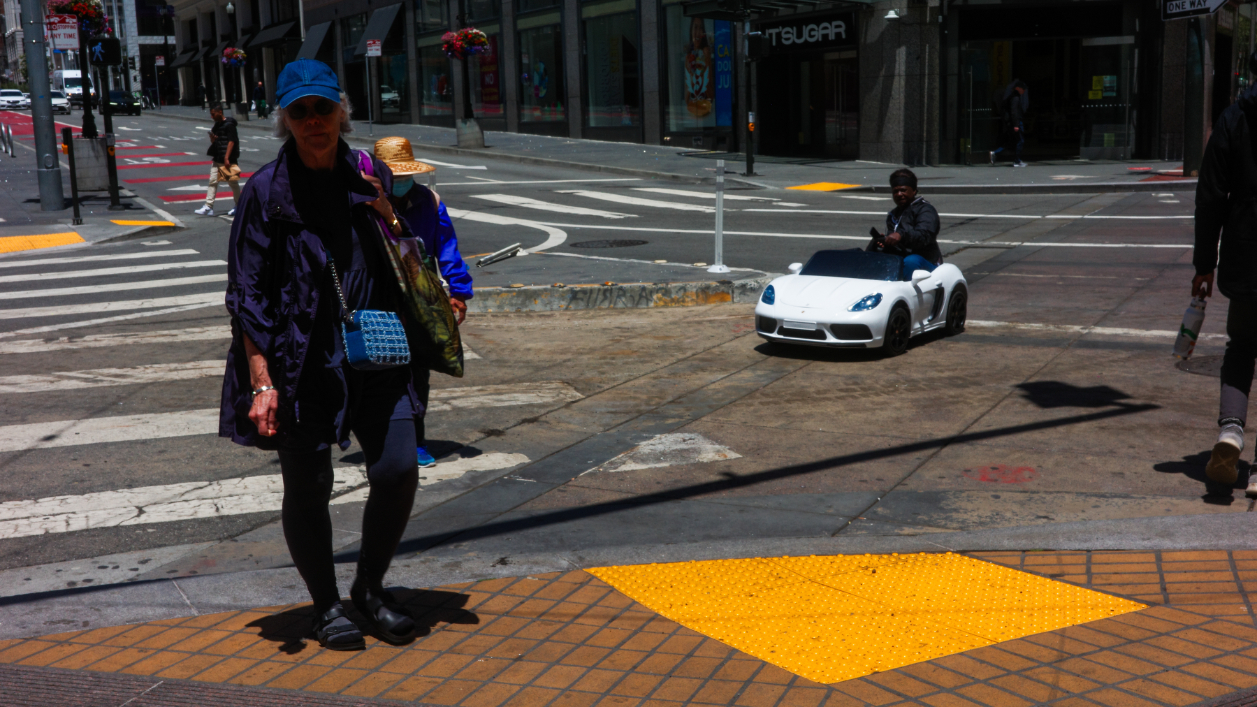 An adult in a toy car crosses an intersection while adults walk nearby.