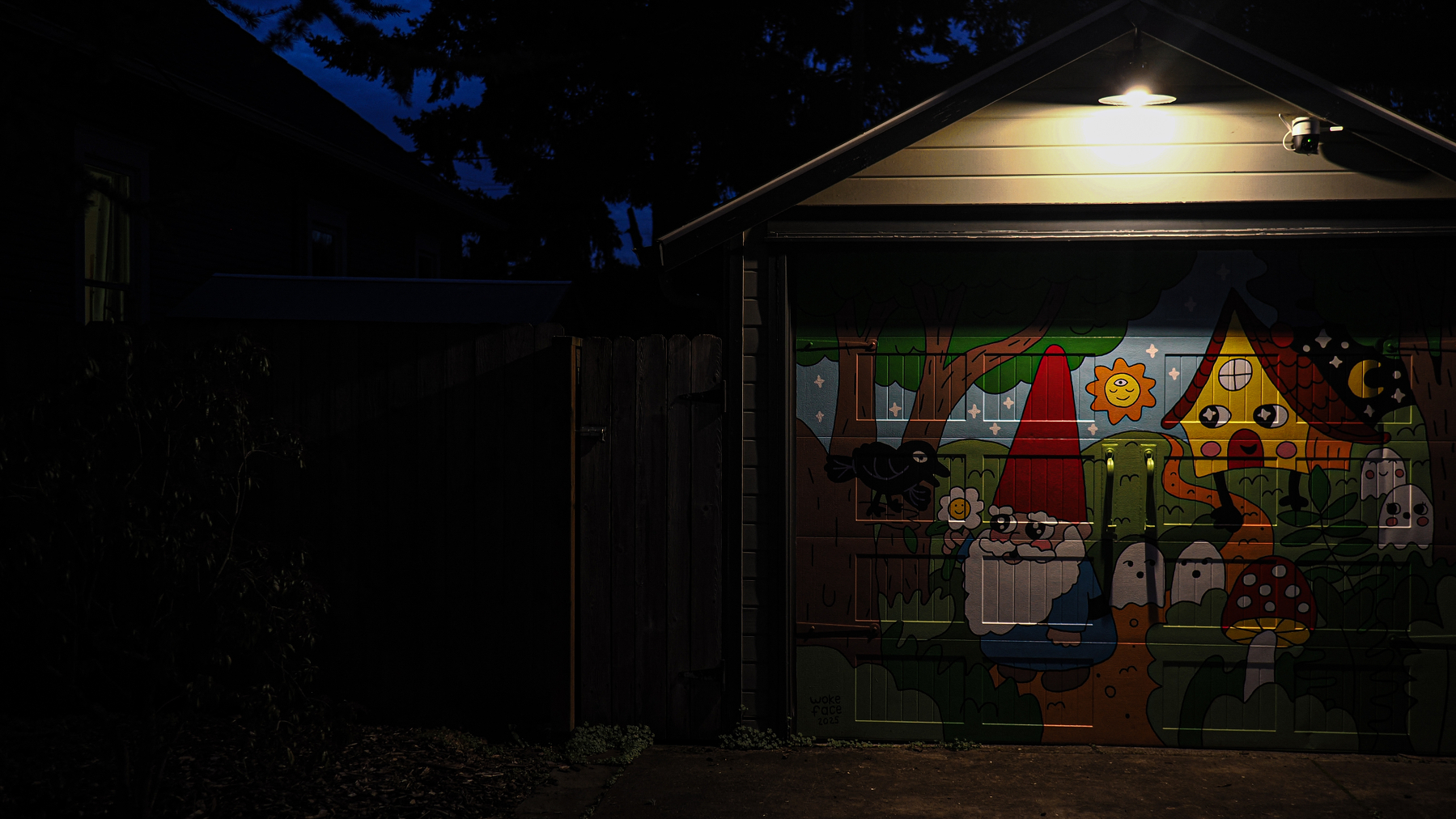 A garage door features a colorful mural of gnomes, mushrooms, and whimsical houses, illuminated by a light above.