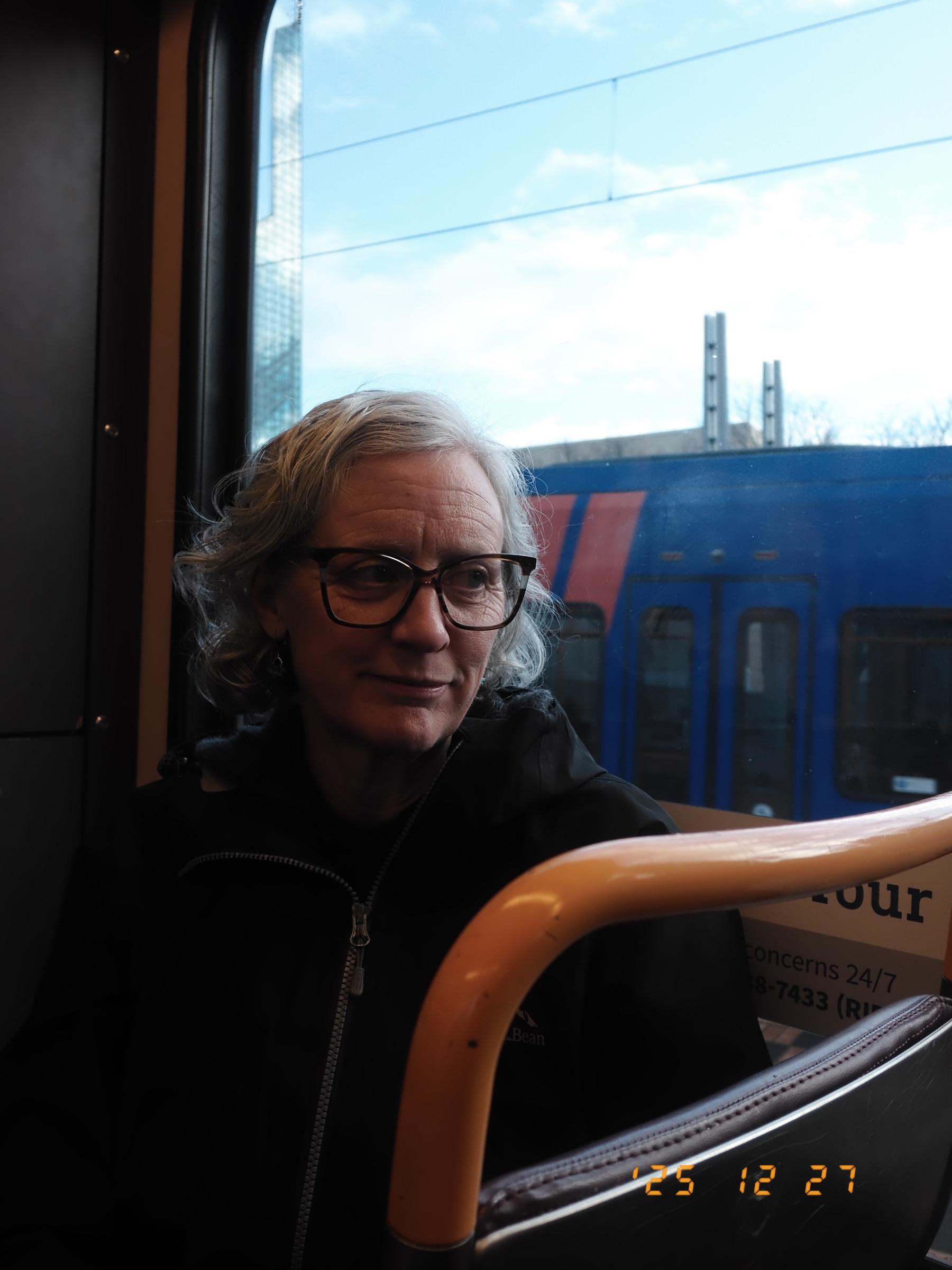 Auto-generated description: A person wearing glasses and a dark jacket is sitting by a window on public transportation, with a blue train visible outside.