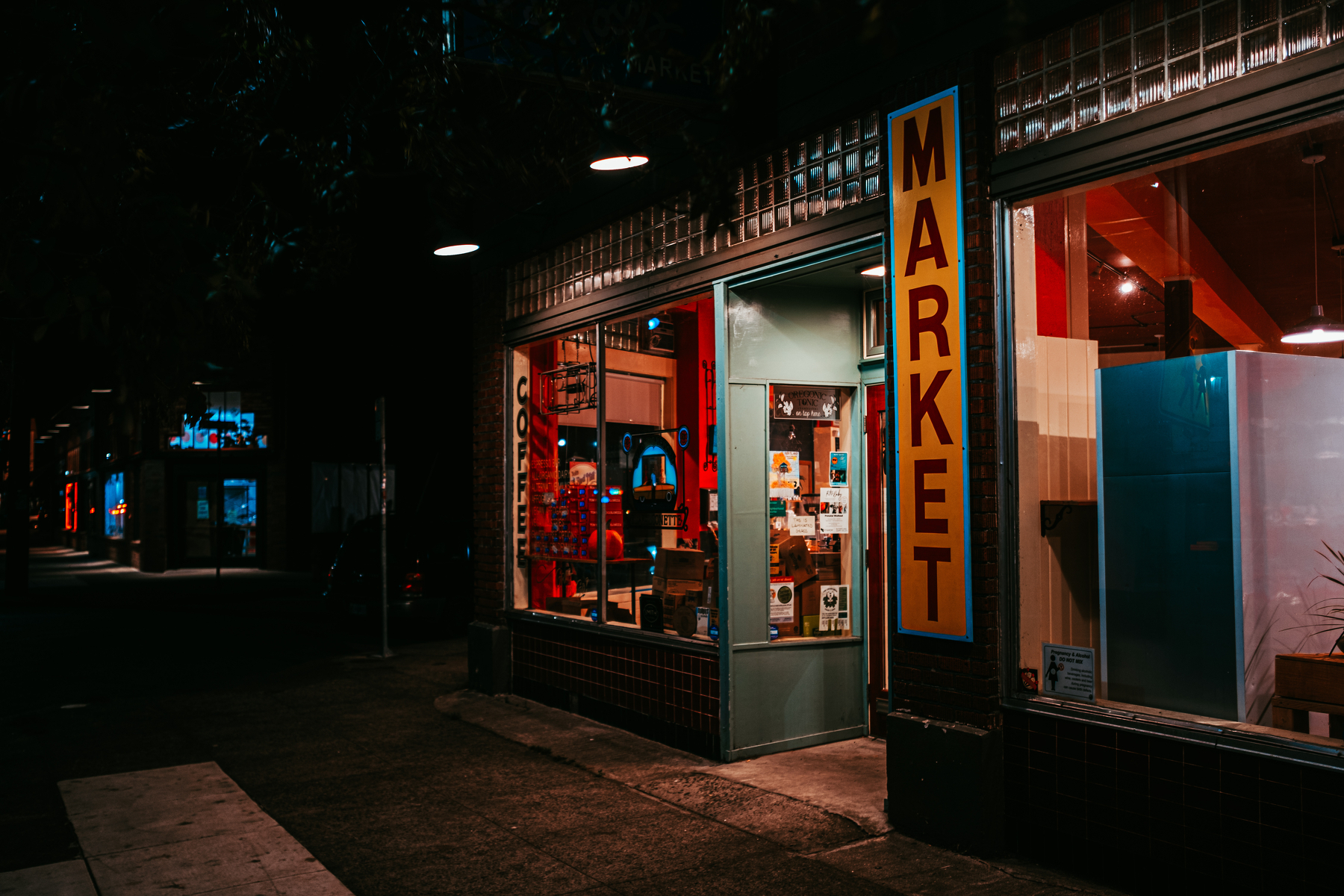 A small market storefront is dimly lit under streetlights at night, with various items visible through its windows.