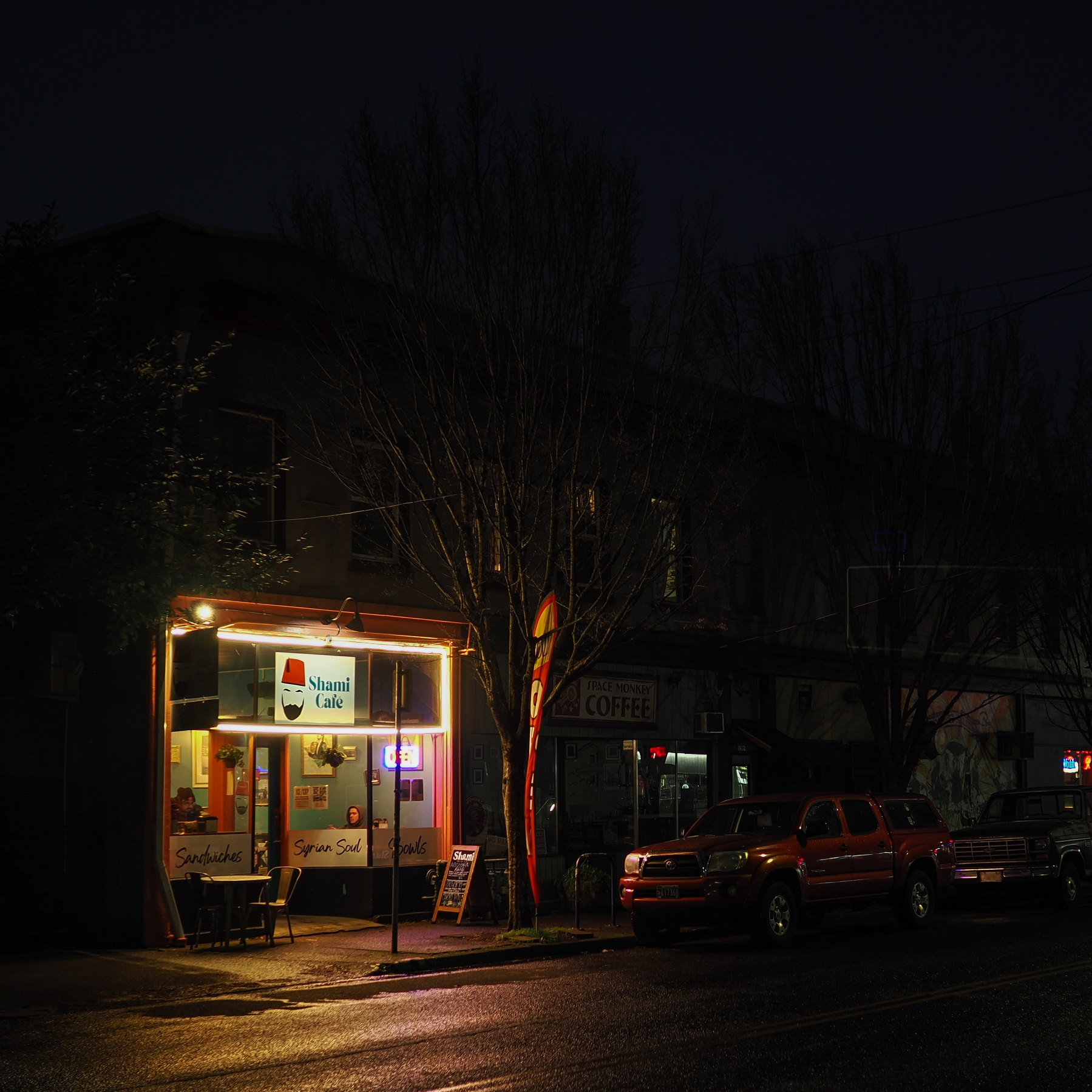 A small, warmly lit café with a sign outside stands on a dark street next to parked cars.