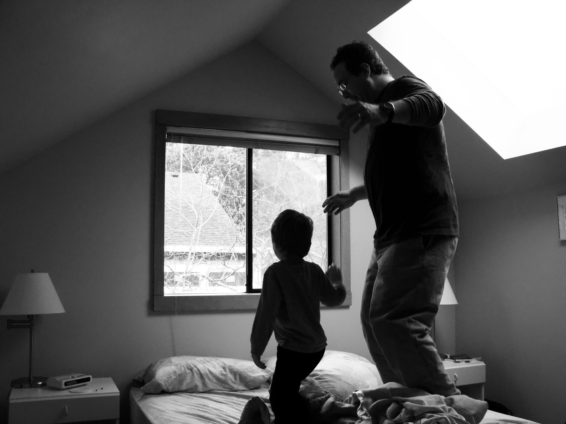 A child and an adult joyfully play and jump on a bed in a softly lit room.
