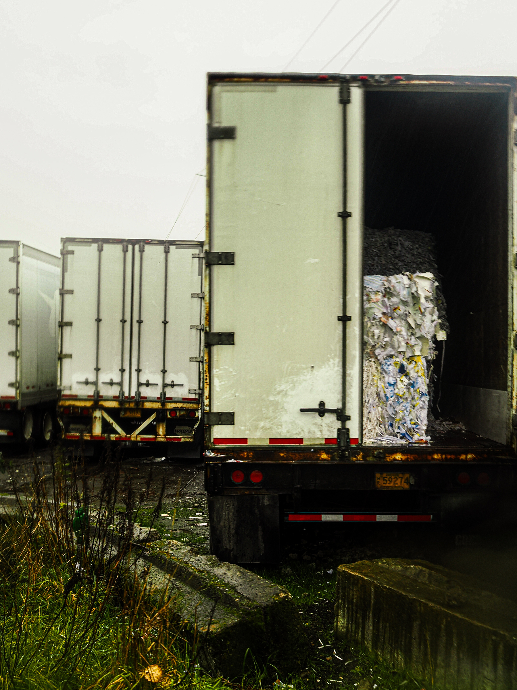 A semi-truck trailer is partially open, revealing baled recyclable materials inside, with other trailers parked nearby in a rain-soaked industrial area.