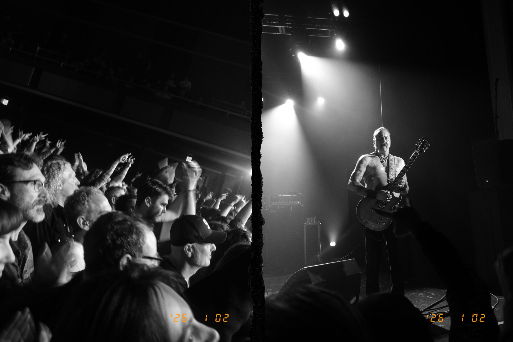 A black and white photo captures a crowd at a concert with a guitarist performing on stage under dramatic lighting.