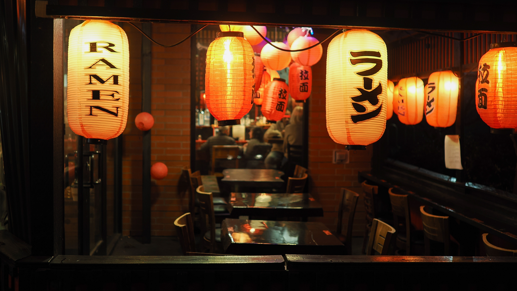 A cozy restaurant interior is warmly illuminated by red and orange lanterns with RAMEN written on them in both English and Japanese.