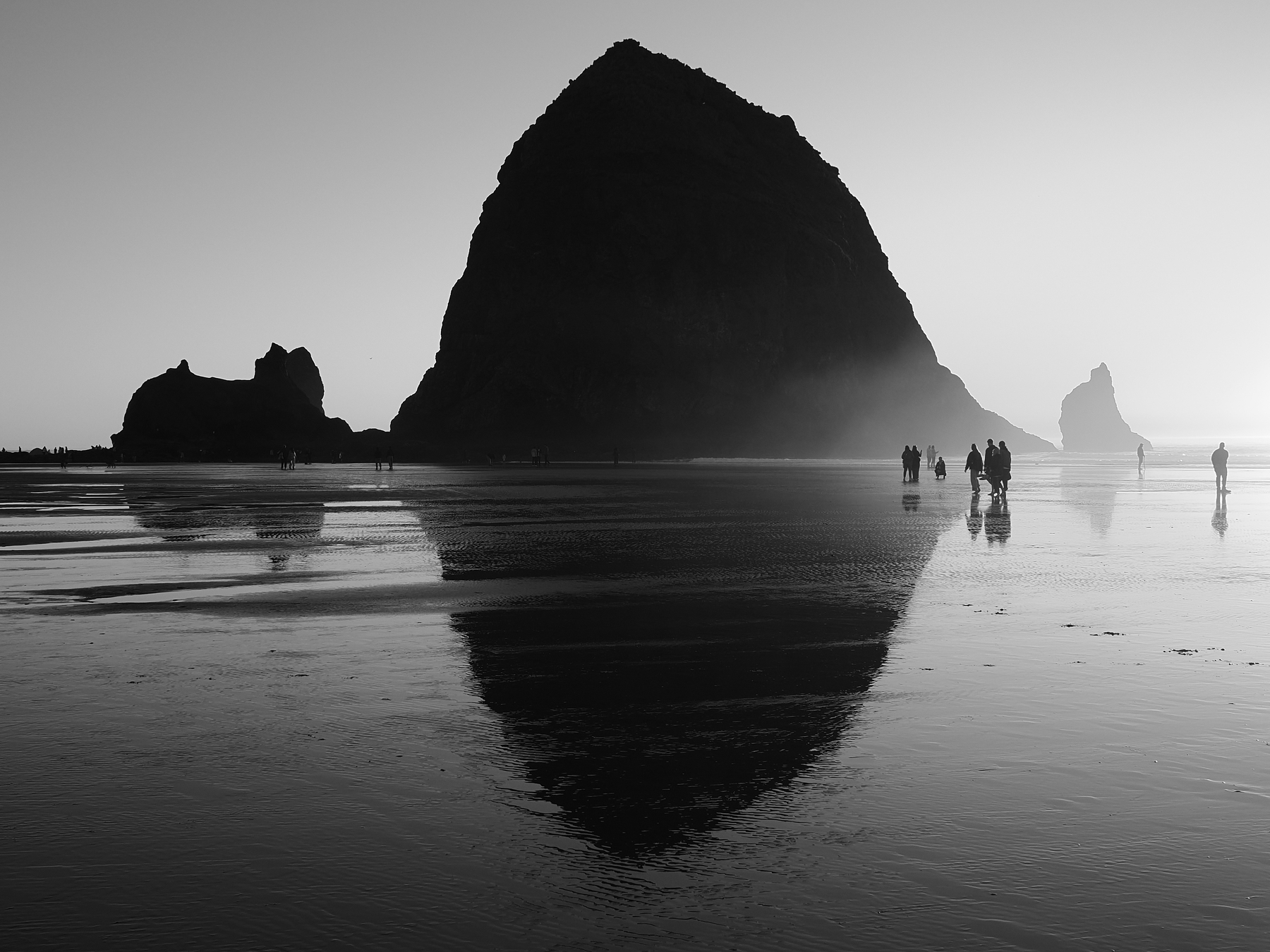 A group of people walk along a reflective beach at sunset with a large rock formation in the background.