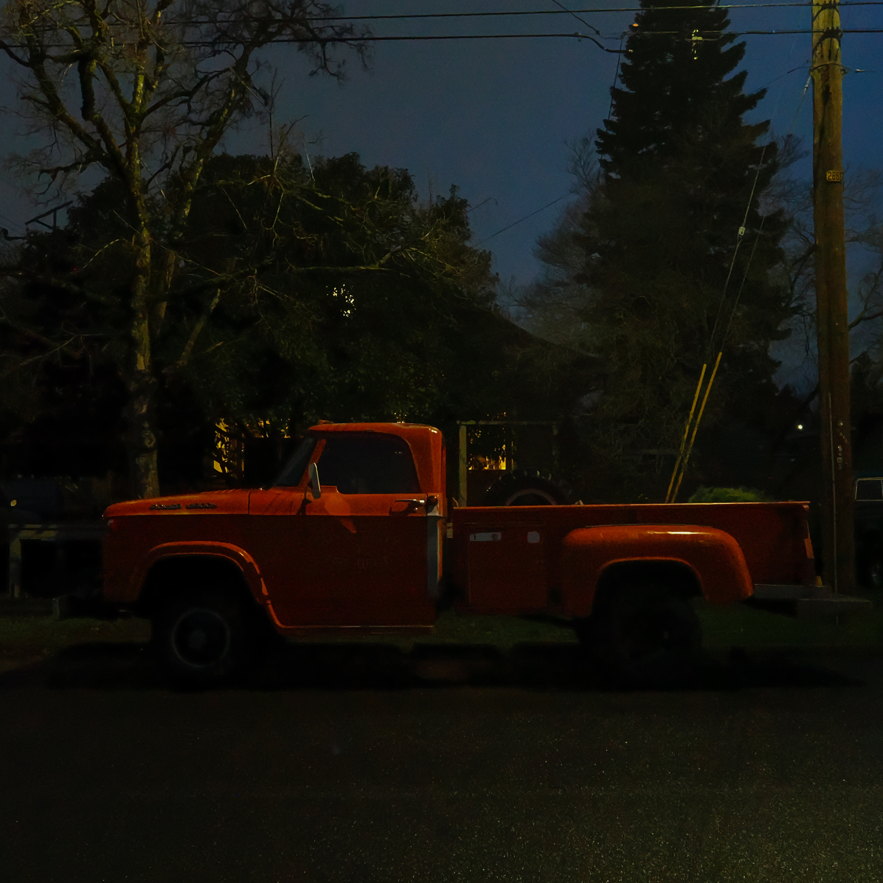 A red truck is parked on a dimly lit street at night, surrounded by trees and utility poles.