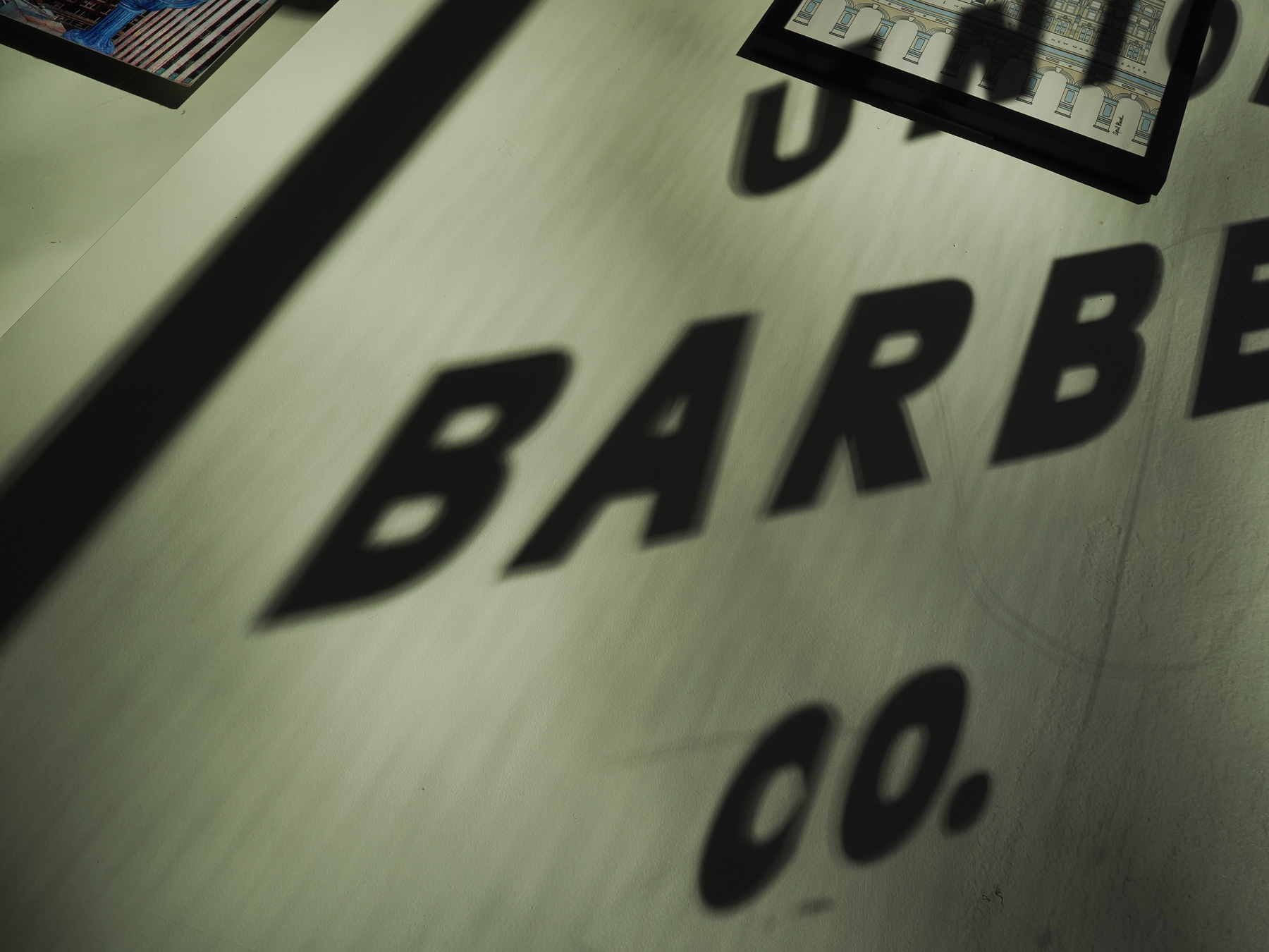 Shadows of the words BARBER CO. are cast on a surface, with partial views of framed pictures in the background.