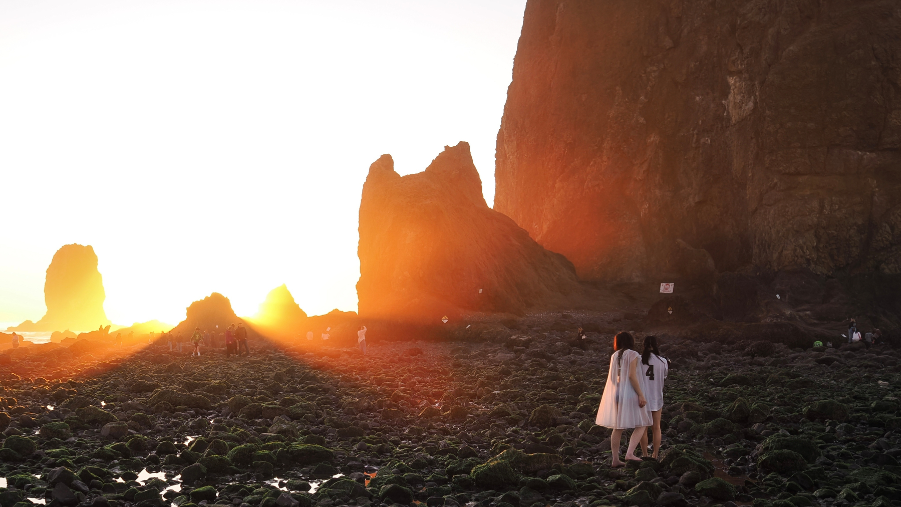 Two people in white dresses walk along a rocky coastline at sunset, with large sea stacks in the background.