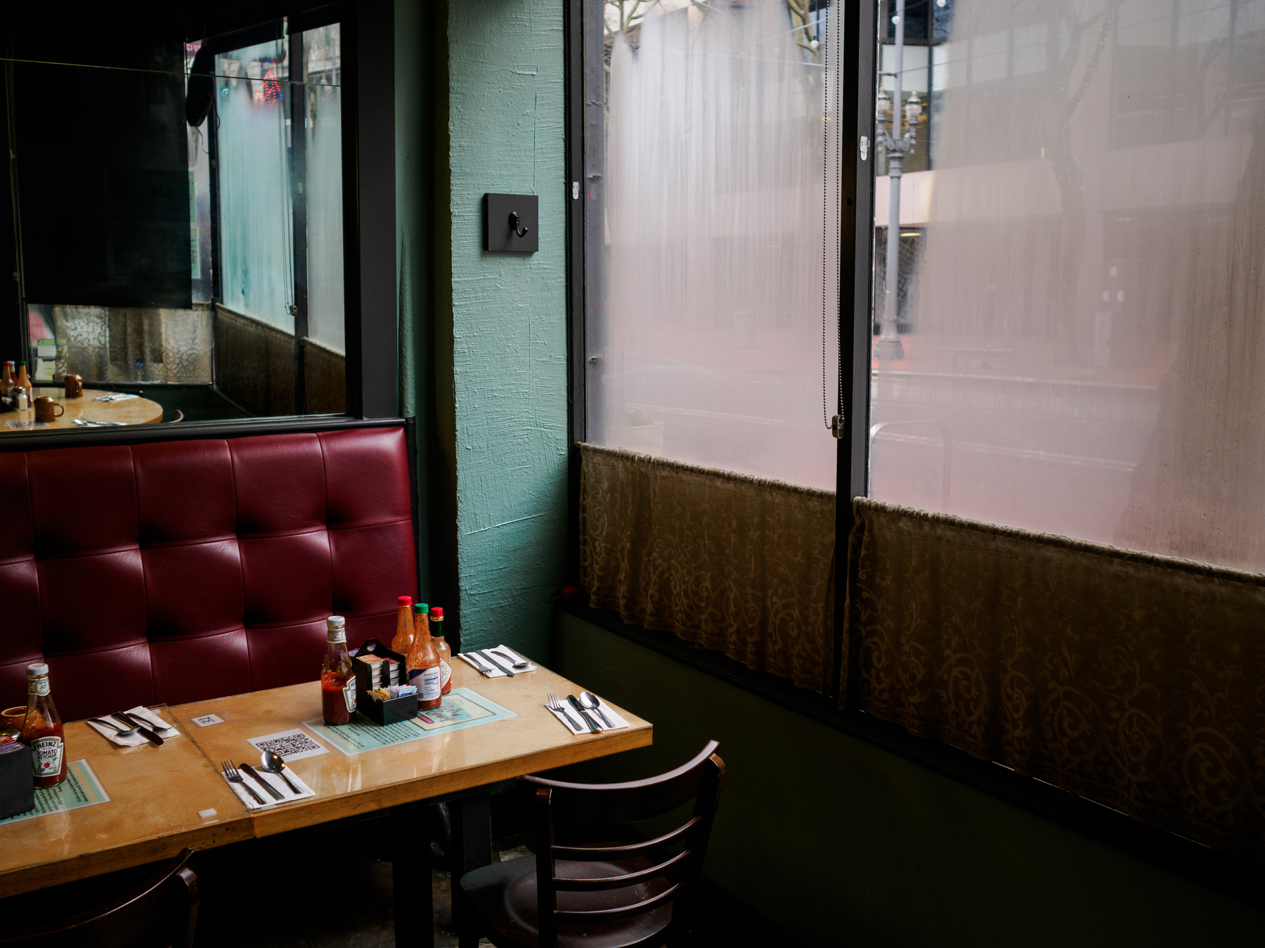 A small restaurant table set for four is next to a window with frosted glass, displaying condiment bottles and neatly arranged silverware.