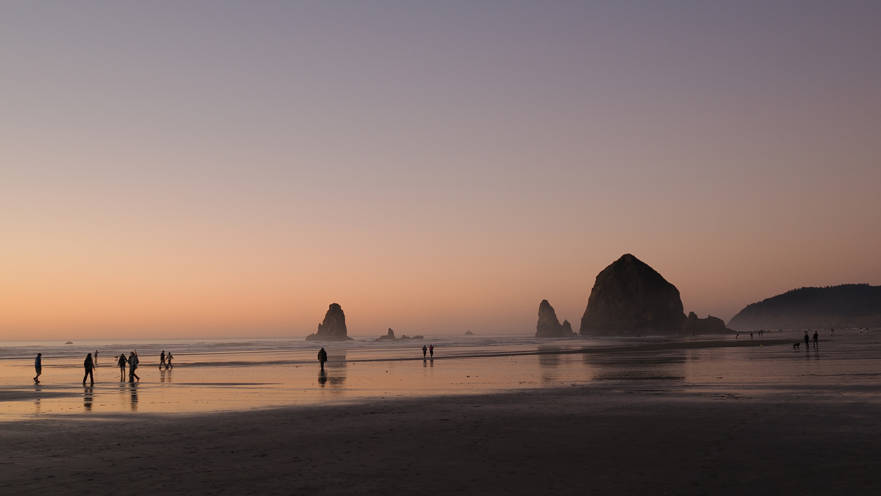 A serene beach scene at sunset features silhouettes of people walking along the shoreline with large rock formations in the background.